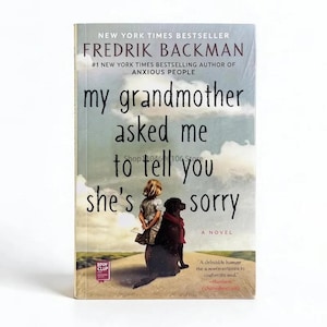 Puede incluir: Un libro de bolsillo titulado My Grandmother Asked Me to Tell You She's Sorry de Fredrik Backman. La portada muestra a una niña y un perro negro en un camino bajo un cielo nublado, con el título y el nombre del autor impresos.