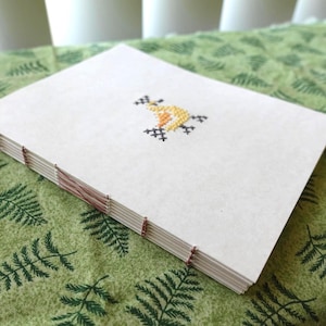 Puede incluir: Un cuaderno rectangular con cubierta de color crema que presenta un pequeño diseño central en punto de cruz de un pájaro amarillo con detalles negros. El lomo está expuesto, mostrando costuras de hilo marrón que unen las hojas de papel blanco.