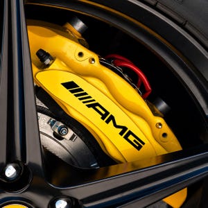 Könnte beinhalten: Ein leuchtend gelber Hochleistungs-Bremssattel mit schwarzem AMG-Logo, montiert hinter einem schwarzen Mehrspeichenrad. Die Baugruppe umfasst eine metallische Bremsscheibe mit einer sichtbaren silbernen Schraube und roten Kabeln.