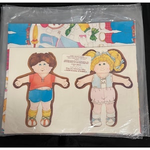May include: Cabbage Patch Kids paper gift wrap sheets in a clear plastic sleeve. The sheets feature two die-cut doll figures, one with brown hair in a red shirt and blue shorts, and one with blonde pigtails in a blue vest and pink bloomers.