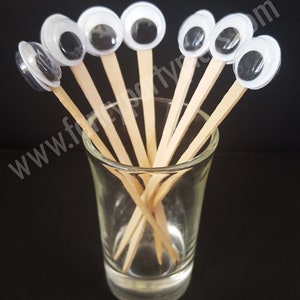May include: A set of eight googly eyes on wooden skewers, arranged in a glass shot glass. The googly eyes are white with black pupils and are surrounded by a clear plastic ring.