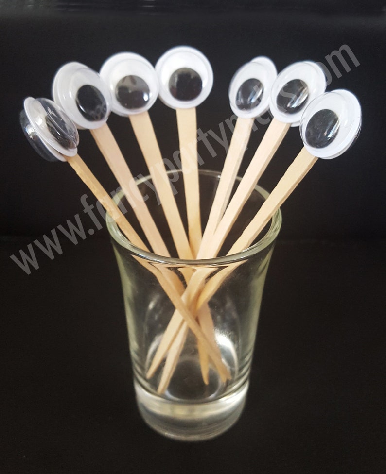 May include: A set of eight googly eyes on wooden skewers, arranged in a glass shot glass. The googly eyes are white with black pupils and are surrounded by a clear plastic ring.