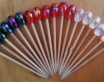 May include: A collection of decorative picks arranged in a fan shape, each featuring a faceted, teardrop-shaped bead at the top. The beads transition in color from dark purple and blue to red, orange, and clear, all attached to wooden sticks.