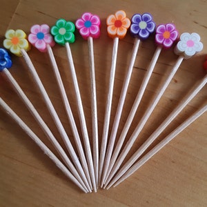 May include: A collection of twelve wooden toothpicks, each topped with a small, colorful flower-shaped topper made of polymer clay. The flowers feature various color combinations, including pink, blue, yellow, green, orange, purple, and white petals.