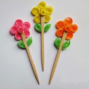 May include: Three paper flowers on wooden sticks. The flowers are yellow, pink, and orange. Each flower has a green leaf on either side of the stem.