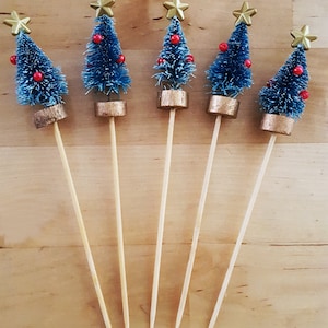 May include: Five miniature Christmas tree decorations. Each tree is blue with red bead ornaments and a gold star topper. The trees are attached to wooden skewers with gold-colored bases, set against a light wood background.