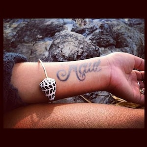 May include: A close-up of a person's wrist with a silver bracelet featuring a black and white shell charm. The word "Agua" is tattooed on the wrist in cursive.