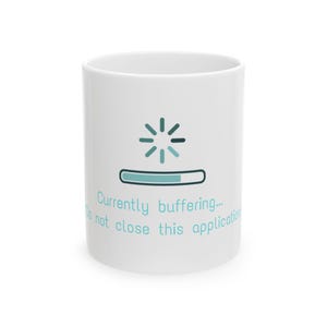May include: A white ceramic mug featuring a teal loading icon and a progress bar graphic. Below the graphic, the text reads: Currently buffering... Do not close this application. The design is centered on the front of the cylindrical mug.