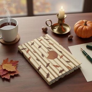 Puede incluir: Un libro de tapa dura con una cubierta de rayas beige y marrones que presenta ilustraciones de hojas de otoño y un gráfico circular central, acompañado de una taza humeante, un candelabro de latón, una calabaza pequeña y dos bolígrafos.