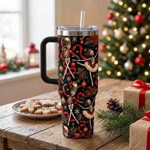 May include: A tall black stainless steel tumbler with a handle and a clear lid featuring a straw. The exterior is decorated with a pattern of dancing skeletons in festive attire, red ribbons, holly leaves, and ornaments on a black background.