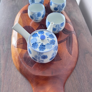 May include: A white ceramic teapot with a side handle and four matching small cups, all decorated with blue floral patterns. The set is arranged on a large, oval-shaped wooden serving tray featuring carved leaf designs and a smooth finish.