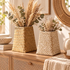 May include: Two trapezoidal woven baskets used as vases, each filled with dried pampas grass, eucalyptus, and small white flowers. The left basket is a natural tan color, while the right basket features a lighter, whitewashed woven texture.