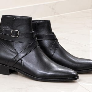 May include: A pair of black leather ankle boots featuring a pointed toe, a low block heel, and a decorative strap that wraps around the ankle, secured with a silver-toned metal buckle on the outer side of each boot for a clean, polished appearance.