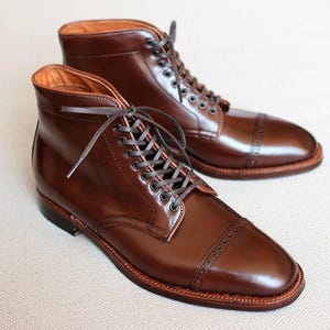 May include: A pair of dark brown leather ankle boots featuring a polished finish, a cap toe with decorative brogue detailing, and a lace-up closure with dark brown laces. The boots have a stacked heel and a visible welt construction along the sole.