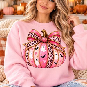 Puede incluir: Una sudadera de cuello redondo rosa claro con un gráfico grande de una calabaza rosa adornada con un lazo de estampado de leopardo. El diseño de la calabaza incluye pequeños fantasmas blancos, murciélagos negros y corazones rosas con detalles de purpurina dorada.