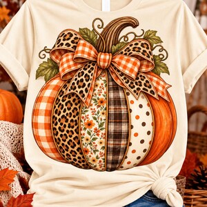 Puede incluir: Una camiseta de color crema con un gráfico grande de una calabaza con segmentos verticales de varios patrones, incluyendo cuadros naranjas, estampado de leopardo, flores, cuadros oscuros y lunares, rematada con un gran lazo decorativo.