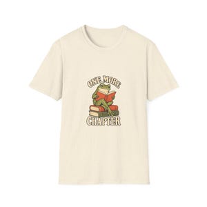 Puede incluir: Una camiseta de manga corta de color crema con un gráfico de una rana verde sentada sobre una pila de dos libros mientras lee un libro rojo abierto. El texto "ONE MORE CHAPTER" está impreso en una fuente curva sobre y bajo la rana.