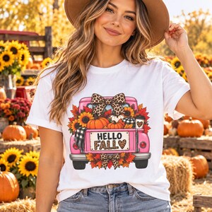 Puede incluir: Una camiseta blanca con el gráfico de una camioneta rosa llena de calabazas naranjas, girasoles y hojas de otoño. La camioneta tiene un lazo con estampado de leopardo y una placa que dice HELLO FALL con un icono de corazón en el diseño.