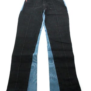 May include: Black denim jeans featuring a high-waisted design with a front button closure. The legs are constructed with contrasting light blue side panels that display a circular, swirling graphic pattern. An eye patch is sewn near the waist.