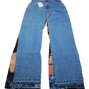 May include: Light blue denim jeans featuring a high-waisted design with a button and zipper fly. The outer side panels of the legs display a dark black and orange tie-dye pattern, while the front and back panels are solid blue denim.