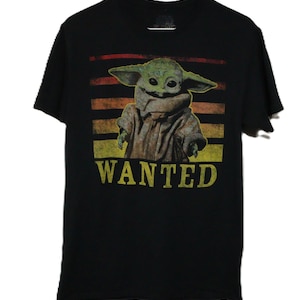 May include: A black short-sleeve t-shirt featuring a graphic of a green creature with large ears and eyes. The graphic is set against a background of horizontal orange and yellow stripes, with the word WANTED printed in yellow capital letters below.