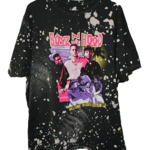 May include: A black short-sleeve t-shirt with a white bleach-splatter pattern. The front features a graphic print with the text "BOYZ N THE HOOD" in pink and "WELCOME TO SOUTH CENTRAL LA" in yellow, depicting a group of people and a car.