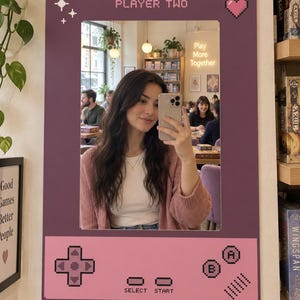 Puede incluir: Un espejo de pared rectangular con un marco de color morado oscuro diseñado para parecerse a una consola de juegos portátil. El marco presenta gráficos pixelados que incluyen el texto PLAYER TWO, un corazón, estrellas, una cruceta y botones A y B.