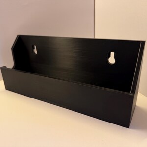 May include: A black rectangular wall-mounted storage shelf with a solid front panel and an open top. The back panel features two integrated keyhole slots for mounting, and the overall construction has a smooth, matte finish.