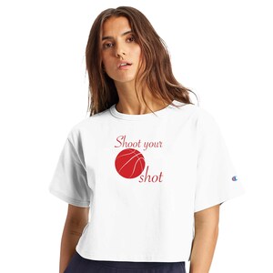 May include: A white short-sleeved cropped t-shirt featuring a red basketball graphic centred on the chest. The text "Shoot your shot" is printed in red script font above and below the basketball. A small blue and red logo is on the left sleeve.