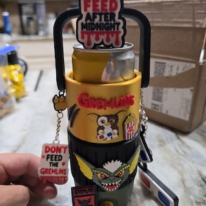May include: A yellow and dark green beverage cooler featuring Gremlins movie imagery. It includes a black handle, a top sign reading DON'T FEED AFTER MIDNIGHT, a Gizmo character, a popcorn bucket, a Stripe character, and a hanging DON'T FEED THE GREMLINS tag.