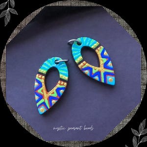 May include: Two handcrafted, teardrop-shaped earrings with a central teardrop cutout. The earrings feature a vibrant design with blue, gold, red, and green geometric patterns. The text "mystic peasant beads" is at the bottom.