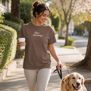 Könnte beinhalten: Ein braunes kurzärmeliges T-Shirt mit dem weißen Schriftzug Slow Mornings auf der Brust, kombiniert mit einer hellen Leggings, dazu ein weißer Einweg-Kaffeebecher mit brauner Manschette und eine schwarze Hundeleine.
