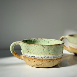 Può includere: Una tazza in ceramica con una forma ampia e arrotondata e un manico. La parte superiore presenta uno smalto verde menta chiaro con macchie scure, mentre la parte inferiore è in argilla beige testurizzata e maculata. Il testo ATOLYE CERAMIC è impresso.