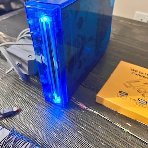 May include: A translucent blue video game console with a glowing blue light at the front disc slot. Included are a grey power adapter, a grey cable, and a yellow box labeled Wii to HDMI Converter with a diagram showing the connection process.