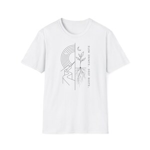 Op de afbeelding: Een wit T-shirt met korte mouwen en een zwarte lijntekening op de borst. Het ontwerp is verticaal gespleten en toont links een zon en een kronkelend pad, rechts een plant met zichtbare wortels en een maansikkel, met de tekst SLOW GROWTH. DEEP ROOTS.