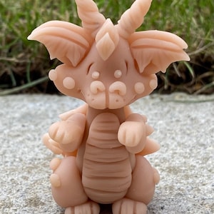 May include: A small, light pink clay dragon figurine with a whimsical expression. The dragon has large, rounded ears, a pointed snout, and a single horn on its head. It has a striped body and small, rounded feet.
