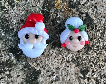 May include: A pair of handmade Christmas ornaments featuring Santa Claus and Mrs. Claus. Santa is wearing a red hat with white trim and a white beard. Mrs. Claus is wearing a white hat with green holly and red berries.