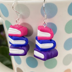 Pode incluir: Um par de brincos de argola de prata com uma pilha de três livros de argila polimérica. Os livros são roxos, azuis e rosados com páginas brancas.