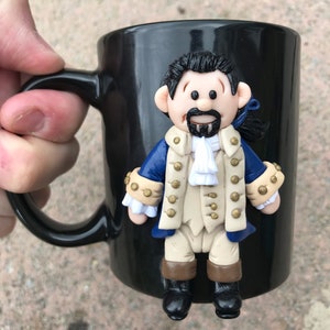 May include: Black ceramic mug with a 3D sculpted figure of a man with a beard and a blue and gold jacket. The figure is attached to the mug.