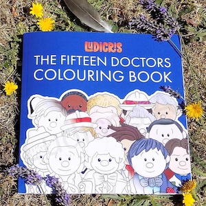 May include: A blue coloring book titled "The Fifteen Doctors Colouring Book" with cartoon illustrations of doctors. The book is surrounded by yellow flowers, lavender, and a feather on a bed of grass.