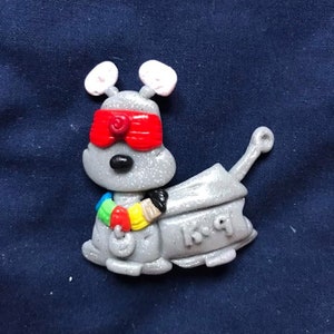 Peut inclure: Une figurine robotique grise et scintillante avec un bandeau rouge, un collier color&eacute; et le texte "K-9" sur le c&ocirc;t&eacute;.