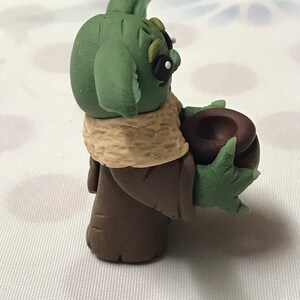 May include: A green and brown hand-crafted figurine of a popular character from the Star Wars franchise. The character is holding a brown bowl in its hand.