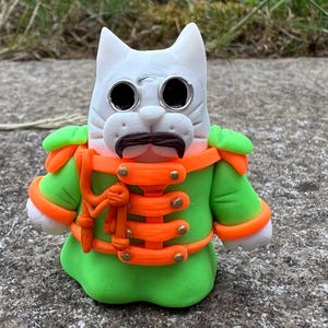 May include: A white cat figurine wearing a green military-style jacket with bright orange trim, gold-colored buttons, and a dark brown mustache. The cat has large, round, silver-rimmed spectacles and a white face with sculpted whiskers.