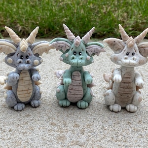 Puede incluir: Tres pequeñas figuritas de dragón de cerámica pintadas a mano. Los dragones son de varios colores, incluyendo gris, verde y blanco. Cada dragón tiene una expresión caprichosa y un cuerpo texturizado y escamoso.