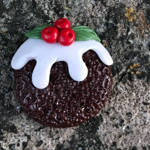 Puede incluir: Un pequeño adorno de pudín de Navidad marrón y blanco con un remolino de glaseado blanco y bayas rojas de acebo.