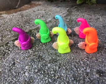 May include: A collection of colorful gnome figurines with vibrant hats in purple, green, blue, pink, neon yellow, and orange. Each gnome has tiny feet and a small bag. The figurines are sitting on a textured, gray surface.