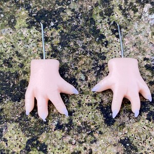 May include: A pair of earrings shaped like hands with long, pointed nails. The hands are a pale pink color and the nails are a light purple color.