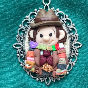 May include: A brown and white figurine of a person wearing a brown hat and a colourful scarf, set in a silver ornate frame.