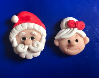 May include: Two handmade Christmas ornaments, one depicting Santa Claus with a white beard and red hat, and the other depicting a woman with white hair and a red bow.