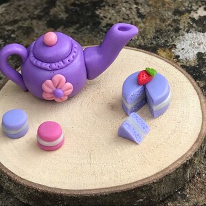 May include: A purple teapot with a pink flower and a pink lid, two purple and pink macarons, and a purple cake with a strawberry on top, all on a wooden slice.